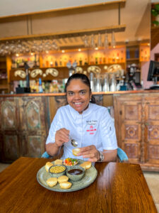 Chef Minal Patel sits at a wooden table eating pani puri, a popular, vegan, Indian street food dish