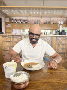 Owner Bobby Patel sits at wooden table in restaurant eating vegan dhal and rice